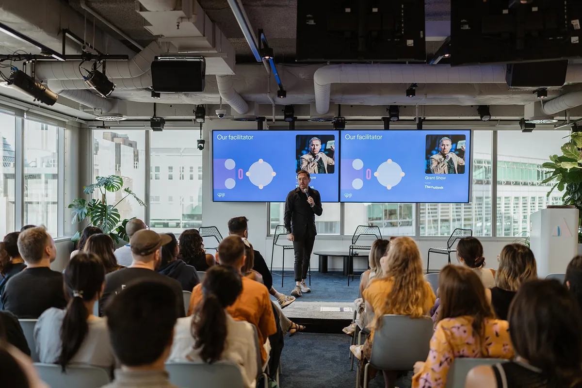 Grant Show presenting on stage to an audience at an Outlook event.